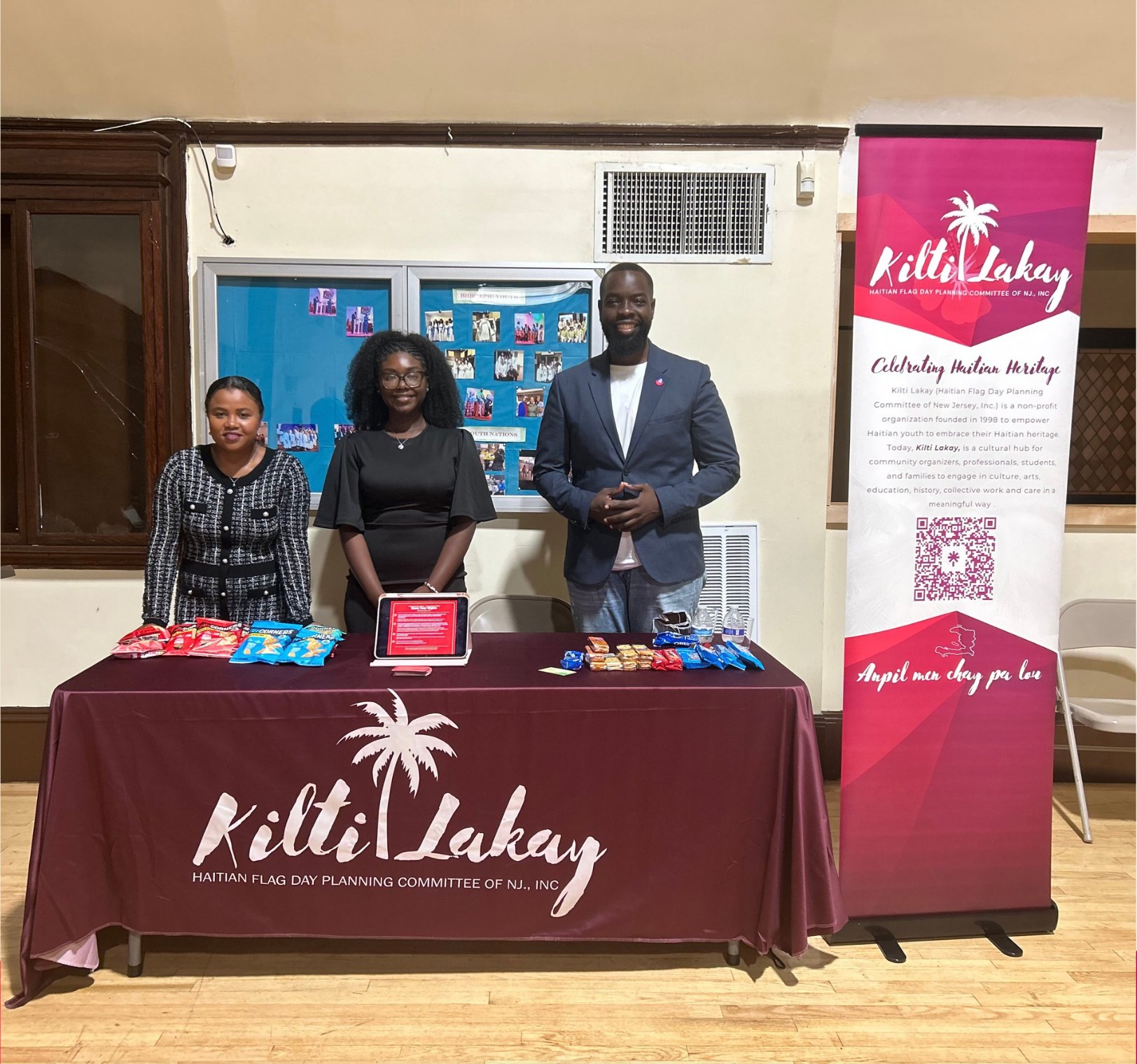 Three Kilti Lakay members standing behind an outreach table with a maroon Kilti Lakay tablecloth, snacks, and a tablet, next to a Kilti Lakay banner in a community hall.