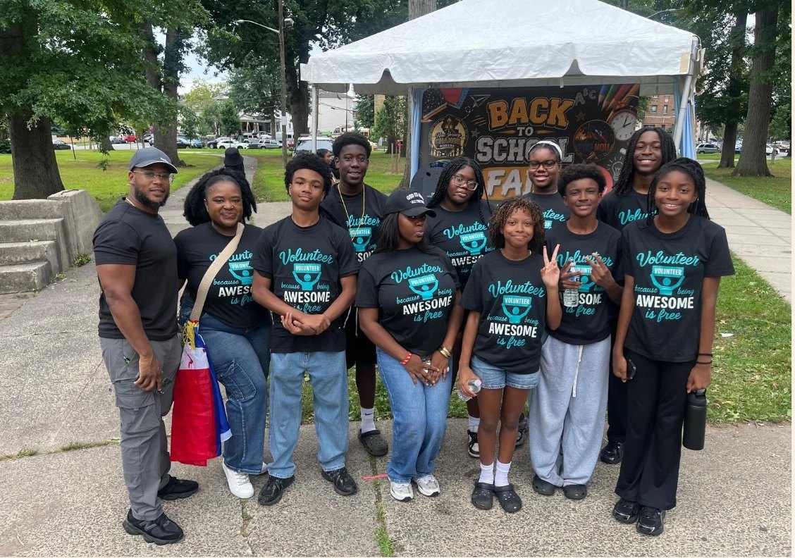 A group of youth volunteers in matching black Volunteer T-shirts gathered under trees at the Back to School Fair tent.